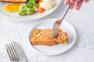 A piece of chicken meatloaf served o the plate with fork and a plate of veggies beside the meatloaf plate