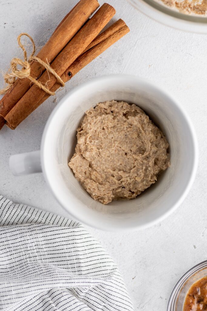 Cinnamon roll dough in a mug with cinnamon sticks besides placed on a table