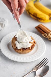 Woman's hand glazing sugar-free cinnamon roll that's on the plate with fork besides and cinnamon sticks and banana in the background, on the table