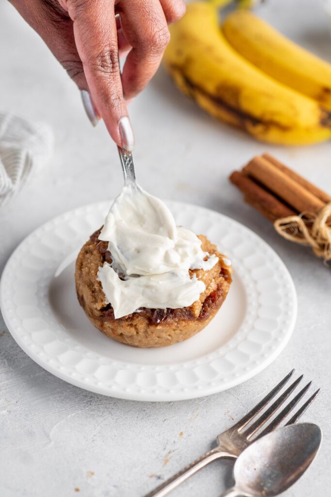 Woman's hand glazing sugar-free cinnamon roll that's on the plate with fork besides and cinnamon sticks and banana in the background, on the table