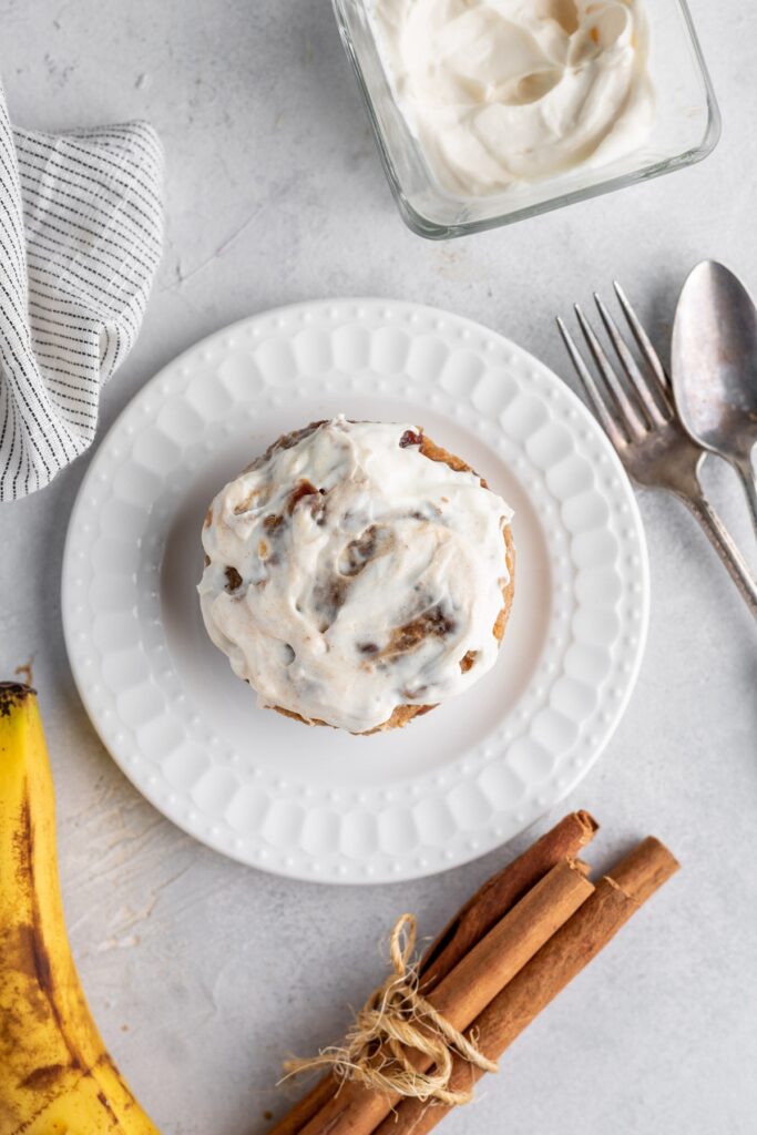Delicious sugar-free cinnamon roll on the plate served on the table with cinnamon sticks, banana, fork, spoon and napkin