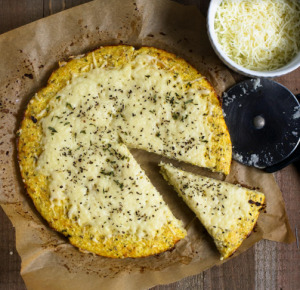cauliflower pizza crust with cheese on a slice on parchment paper and a bowl of extra cheese