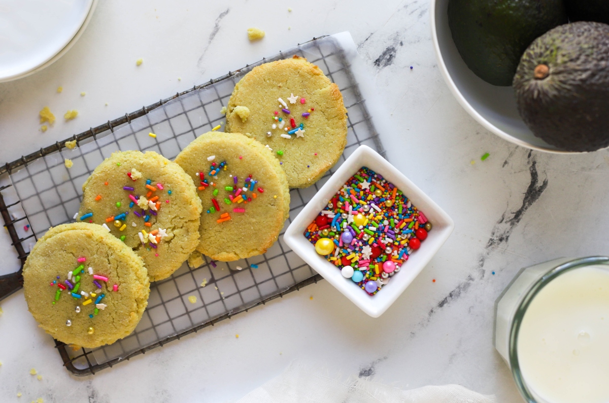 Keto sugar cookies with a cup of sprinkles, a bowl wit avocadoes and a bottle of milk beside