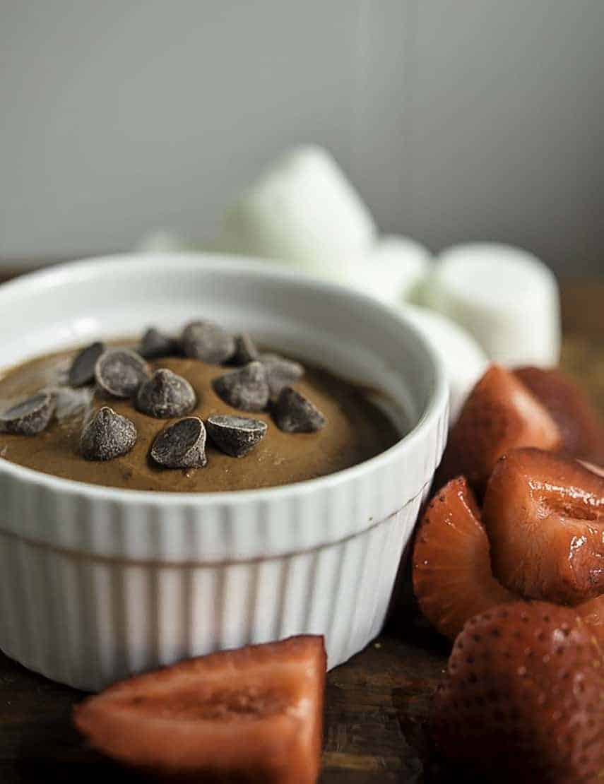 Brownie Dough Dip in a cup sprinkled with chocolate chips, with cut strawberries beside the cup and blurred marshmallows in the background