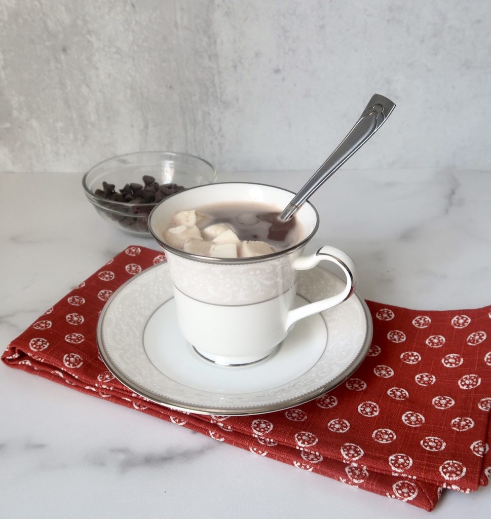 A glass of keto hot chocolate with low sugar marshmallows in in a white mug with a spoon placed on top of a red towel with chocolate chips in the background