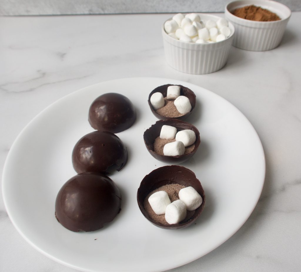 6 chocolate shells on a white plate with half of them filled with marshmallows and cocoa mix. two small bowls of ingredients in the background