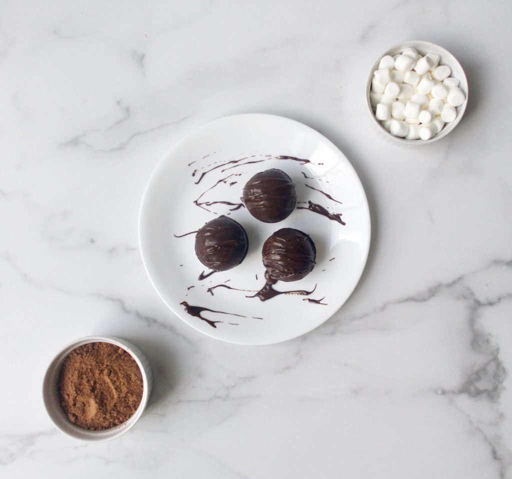 3 chocolate bombs on a white plate with a bowl of marshmallows and a bowl of hot cocoa mix