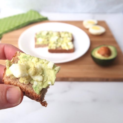 Avocado egg salad spread on sliced toast with hand holding a bite sized piece up to camera