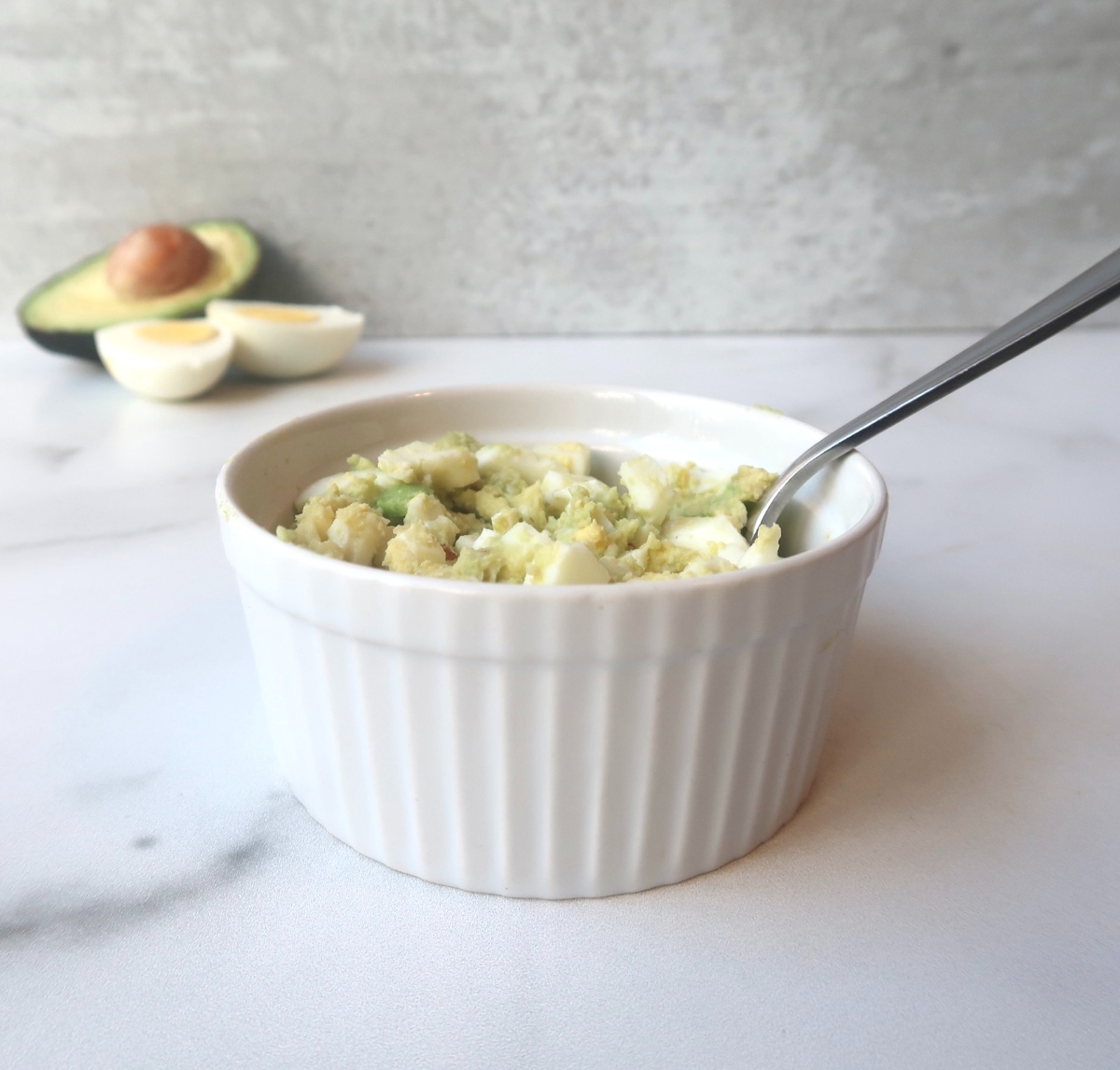 white ramekin filled with avocado egg salad and a spoon with avocado and hard boiled egg in background 
