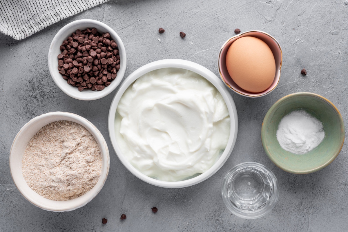 Food ingredients for Greek yogurt pancakes: Greek yogurt in bowl, chocolate nibs in small bawl, egg, flour in bowl, water in glass and baking soda