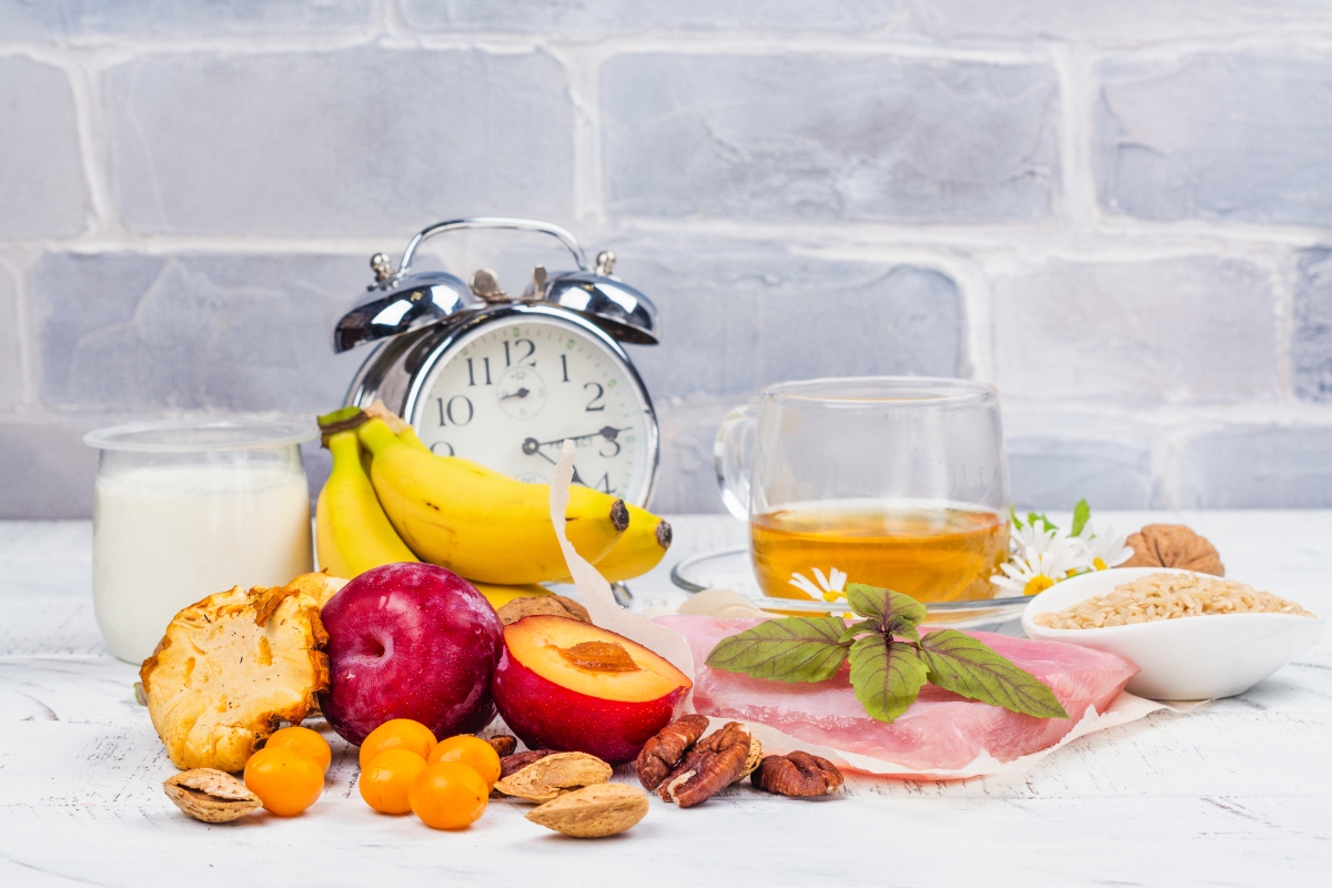 Clock with glass of tea, glass of yogurt, bananas, plums, other fruits, nuts and bowl of seeds on table