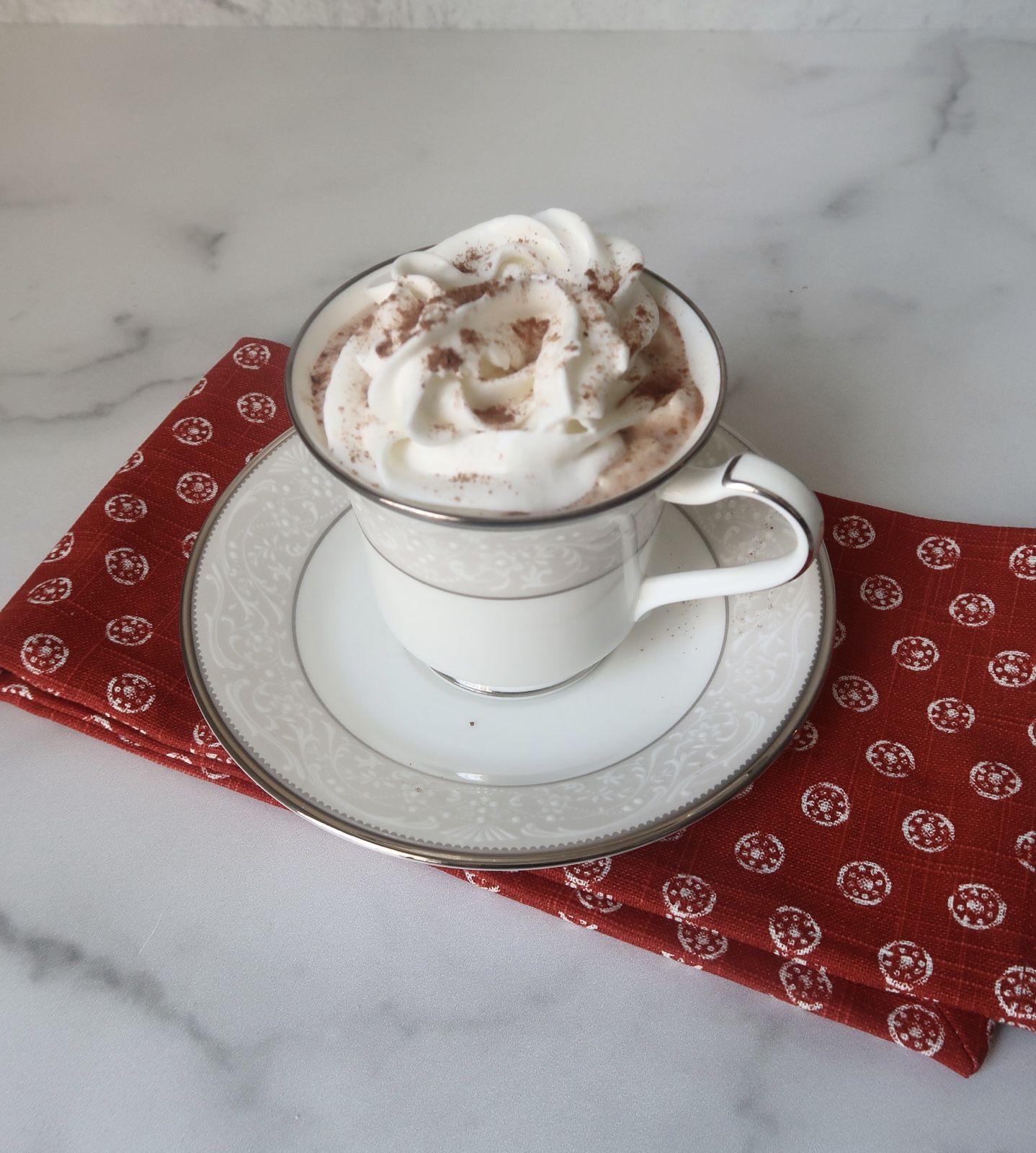 A white mug filled with low carb hot chocolate with whipped topping sits of a red towel on a marble countertop