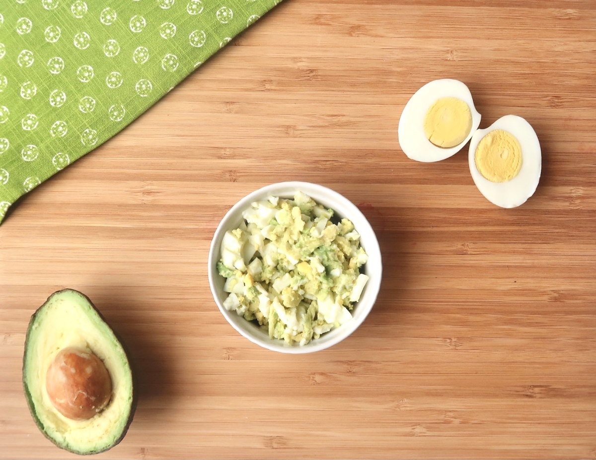 Avocado egg salad in white bowl next to sliced avocado and sliced hard boiled egg