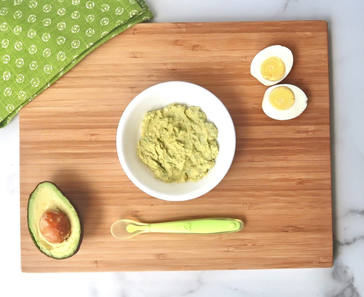 mashed avocado egg salad in white bowl with baby spoon, sliced avocado half, and sliced hard boiled egg