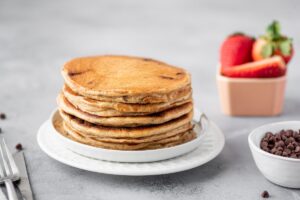Greek yogurt pancakes pilled up together on a plate with bowl of strawberries in the background, served on the table