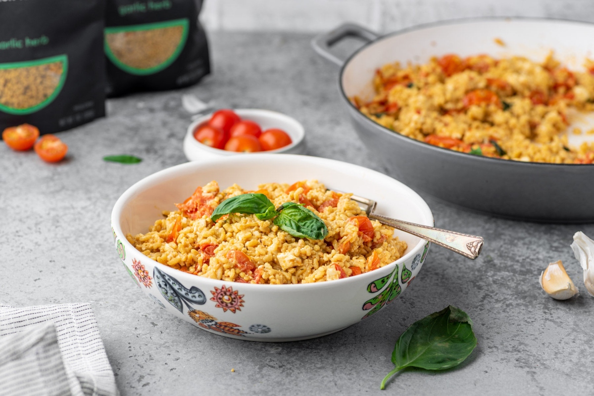 Bowl of low carb feta rice with fork in a bowl and cooking pan with the food, bowl of cherry tomatoes and pack of rice in the background