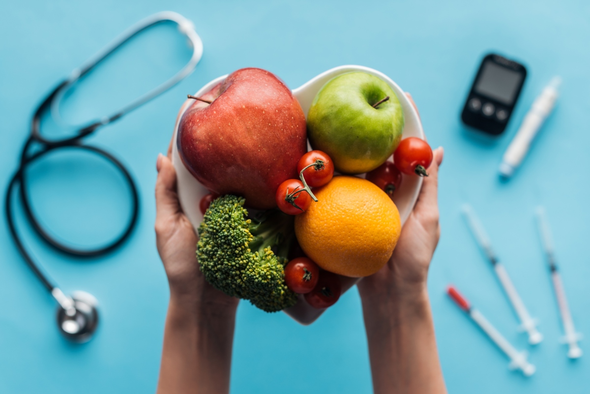 Hand holding heart-shaped bowl of fruits and veggies with blood sugar meter, stethoscope and syringes