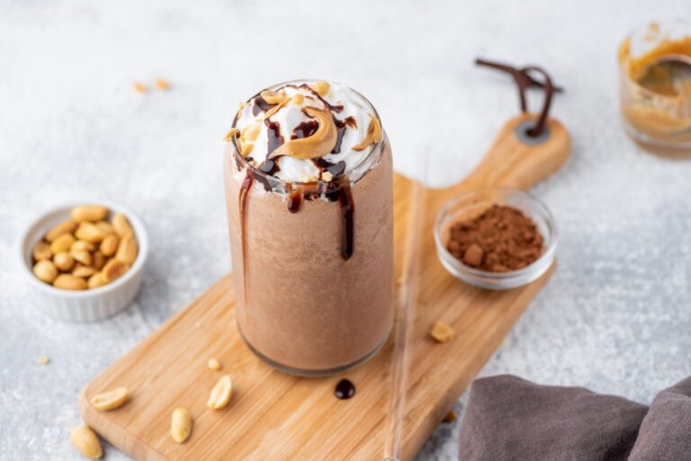 Keto Milkshake toped with cream, chocolate and peanut butter on the cutting board with straw beside and with a cup of cocoa powder beside and peanuts sprinkled around