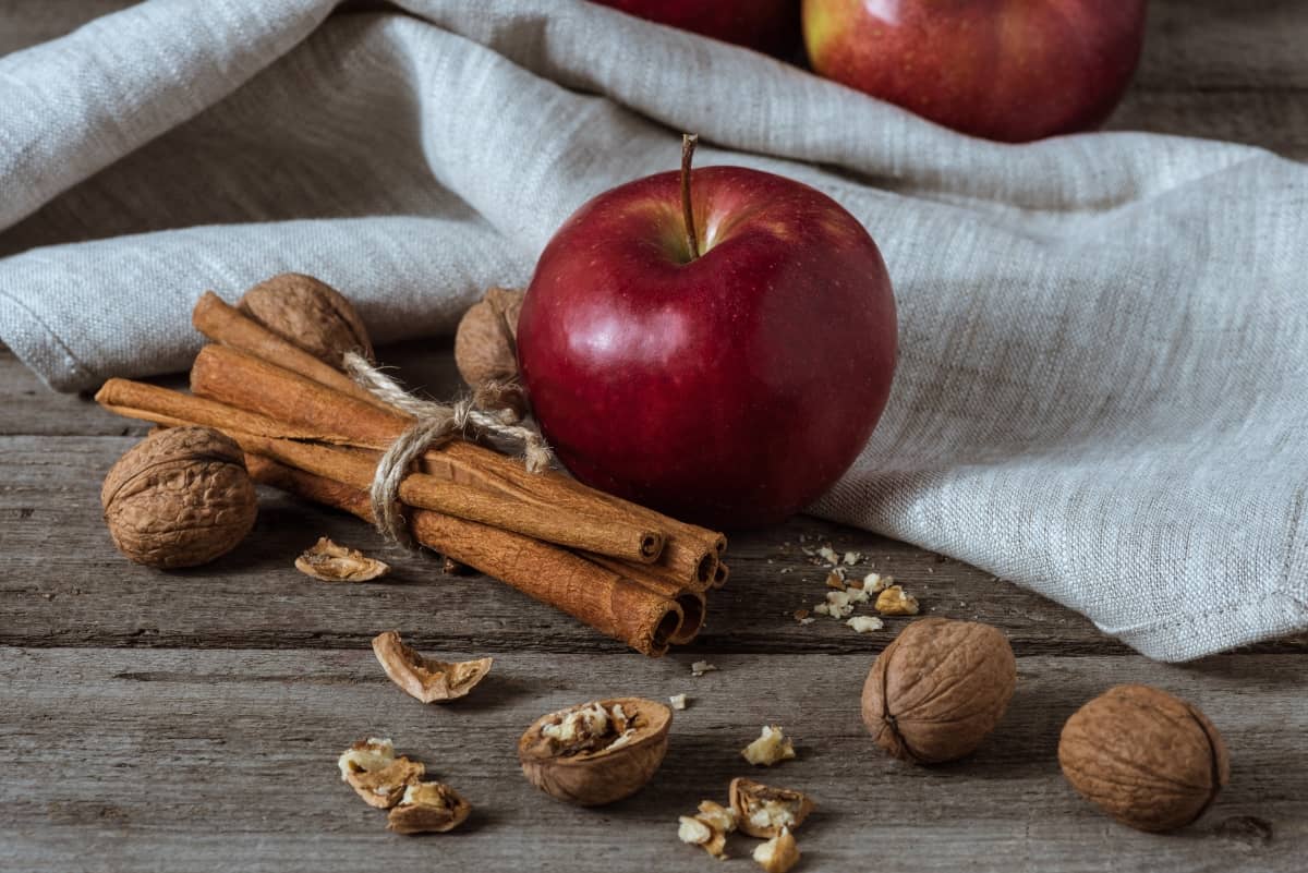 Red apple with cinnamons sticks beside and walnuts with and without shell sprinkled around, with a white cloth in the background on wooden surface