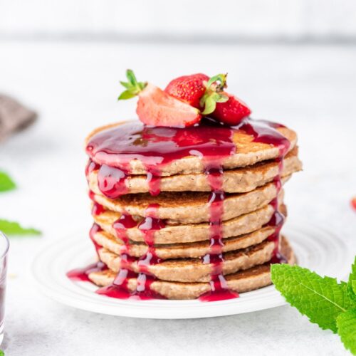 Peanut butter jelly pancakes stacked together with a couple of sliced strawberries on top, served on a white plate and glass cup with jelly and fresh mint leaves on the other side of the plate