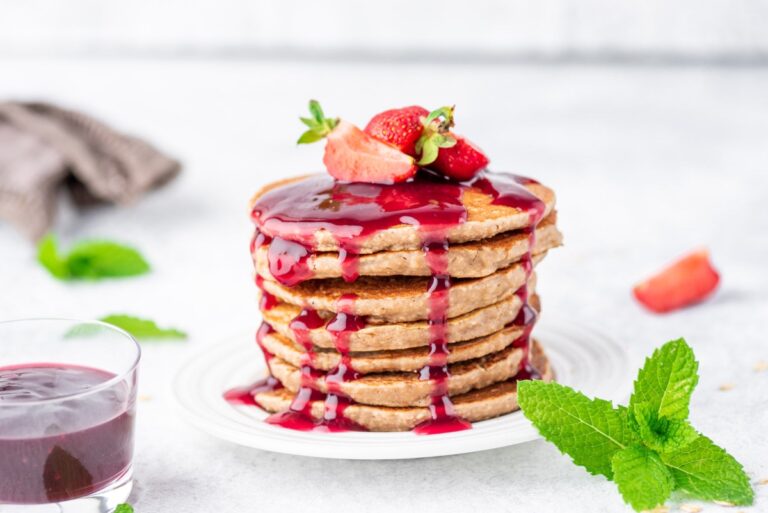 Peanut butter jelly pancakes stacked together with a couple of sliced strawberries on top, served on a white plate and glass cup with jelly and fresh mint leaves on the other side of the plate