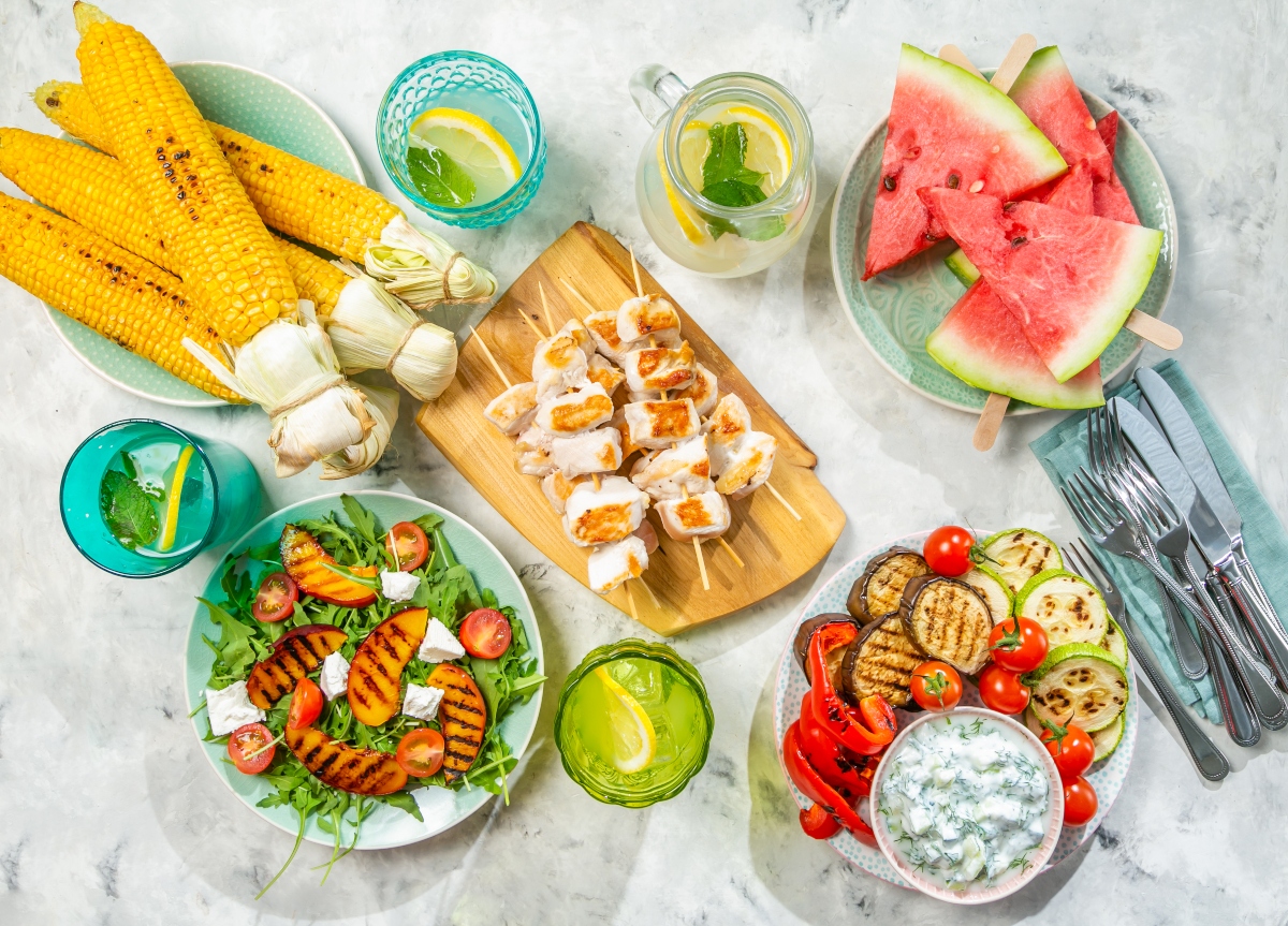 Whole foods on the table, corn cobs, watermelon, grilled vegetables, fruit and cheese salad, chicken on cutting board and glasses of lemonade