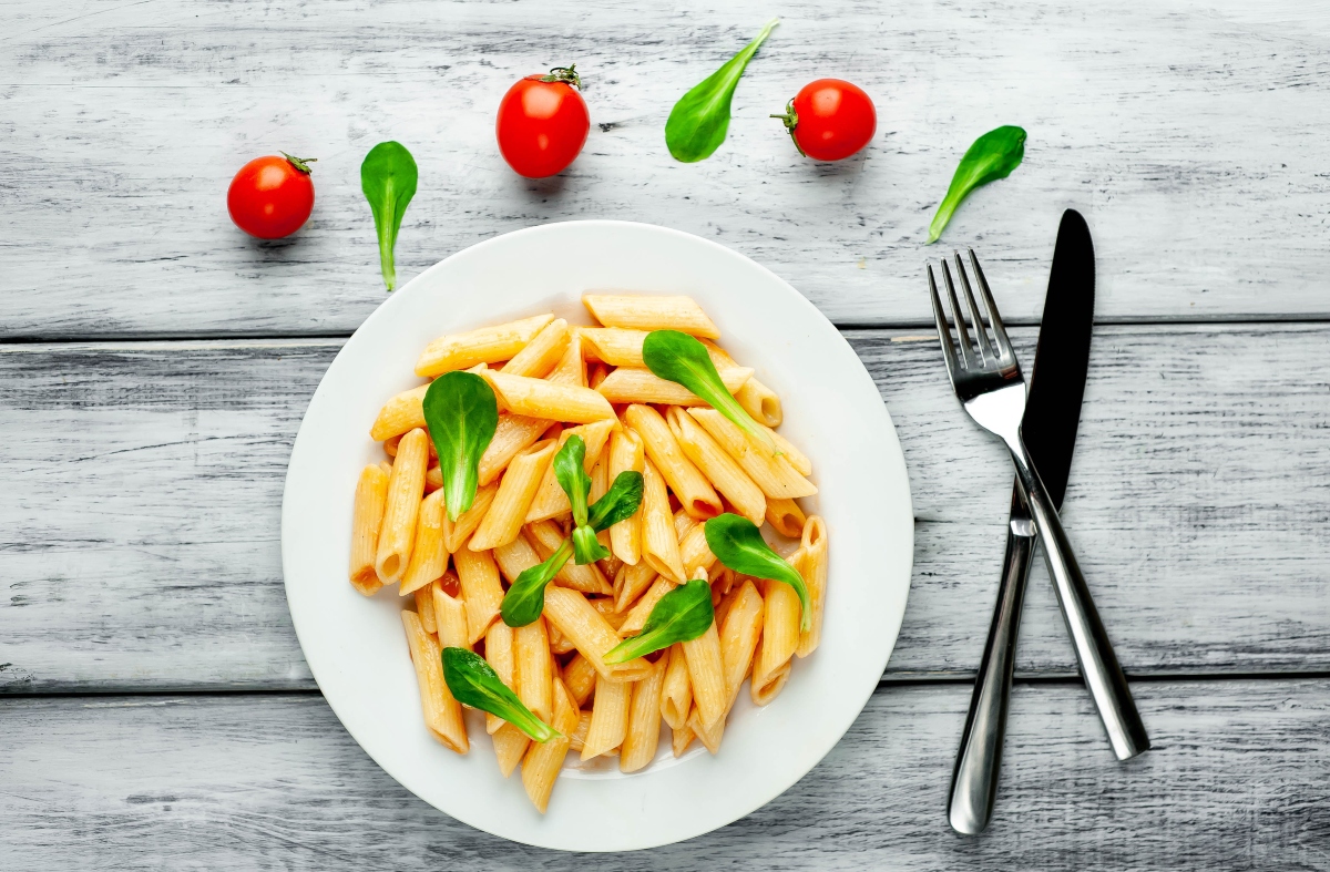 High fiber pasta in a plate with fork and knife beside and cherry tomatoes and basil leaves sprinkled beside the plate