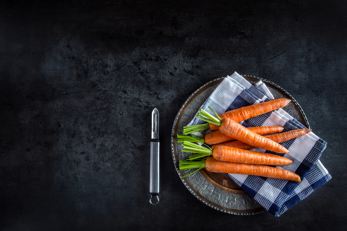 Raw carrots and cloth napkin on a metal plate with a vegetable peeler beside