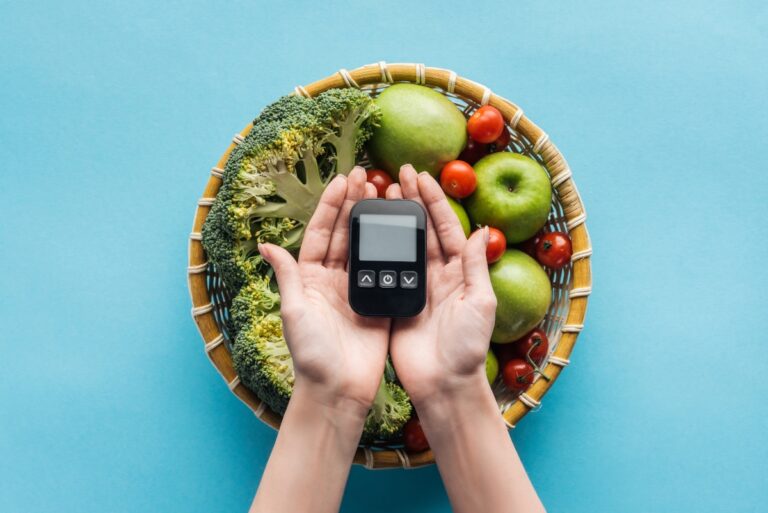 A person holding a glucometer in hands over the bowl filled with vegetables and fruits against the blue background