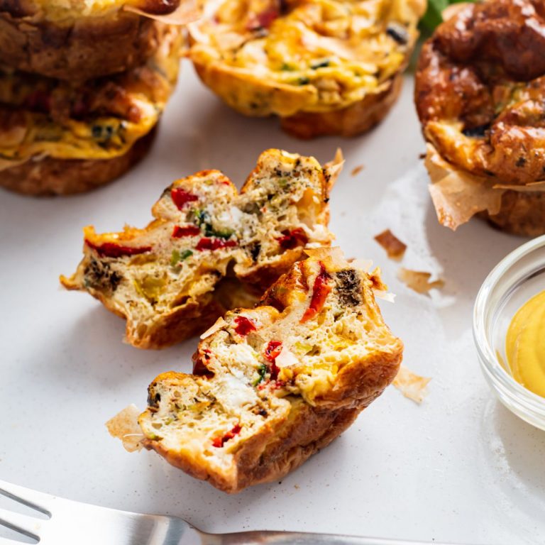 a white plate with several mini quiches. One quiche is cut open to see the vegetables and baked egg inside—a small dish of yellow dipping sauce on the side
