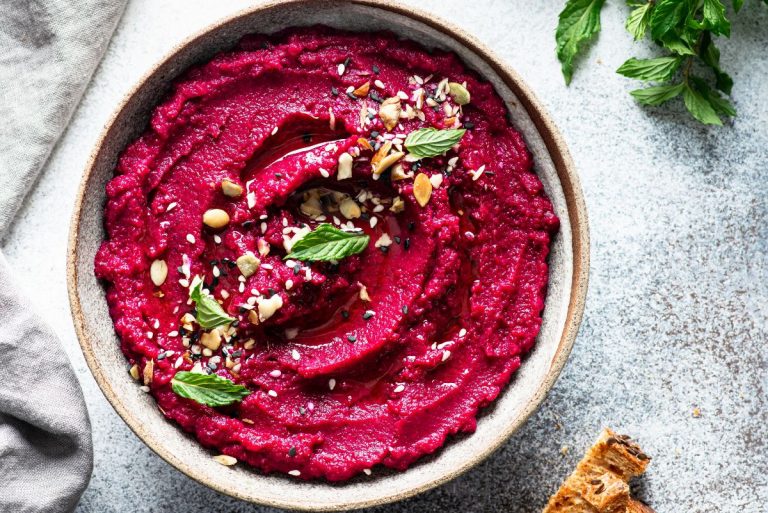 A bowl of beet hummus topped with seeds and herbs on a stone background