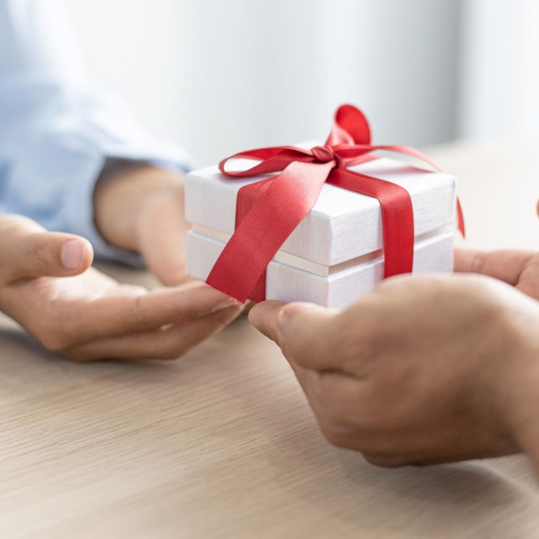 hands of one person giving a gift in a white box with a red ribbon to another person