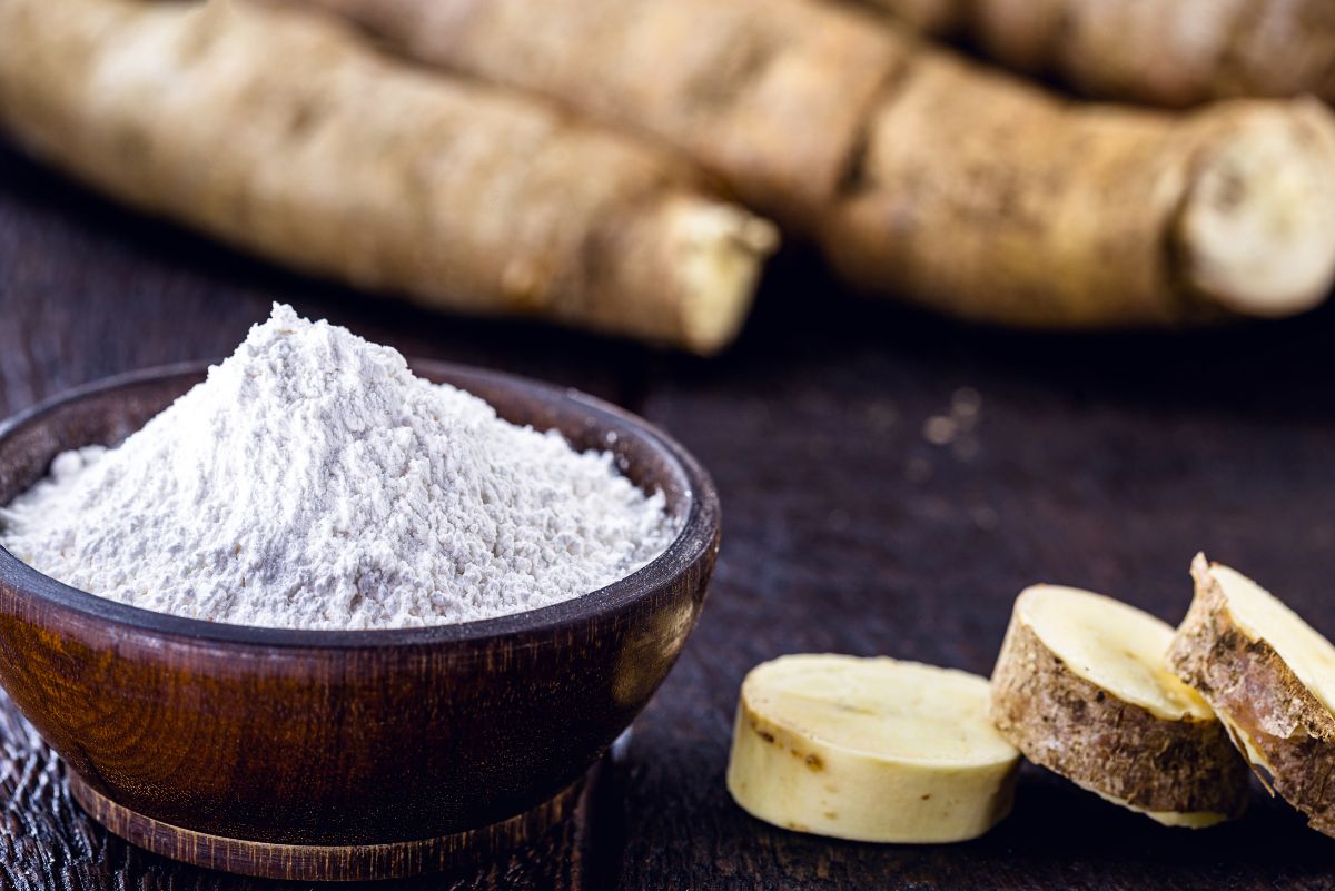 Cassava flour in a wooden bowl and three pieces of chopped cassava root beside the bowl