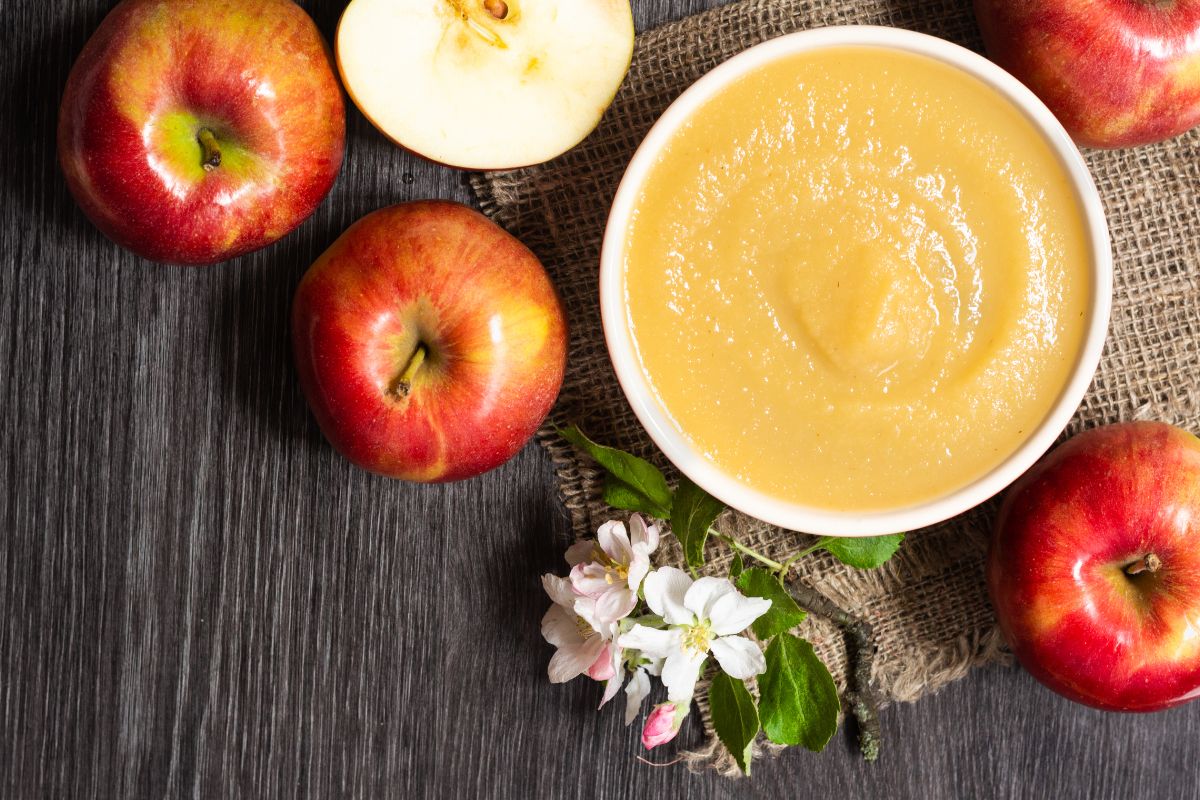 Applesauce in a white bowl and a couple of apples and a flower beside