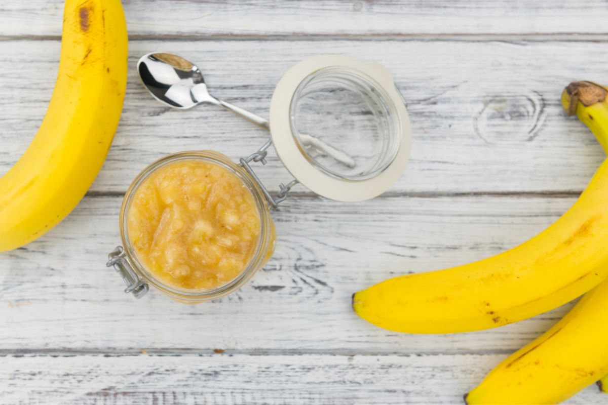 Mashed bananas in a jar with a few bananas and a spoon beside on a wooden surface
