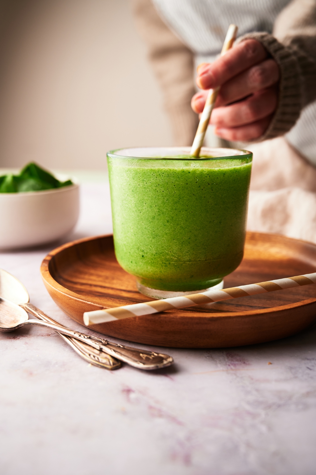 Mango spinach smoothie in a glass with a straw on a wooden tray and a person holding a straw