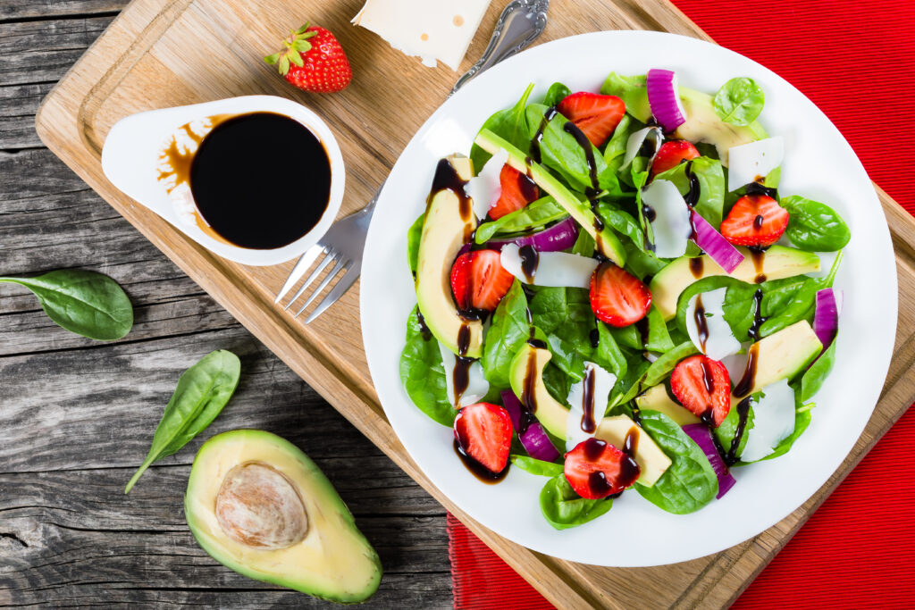 Strawberry,  baby spinach, red onion  goat cheese and avocado salad on a white dish on a cutting board with caramelized balsamic vinegar in a gravy boat, top view, studio lights