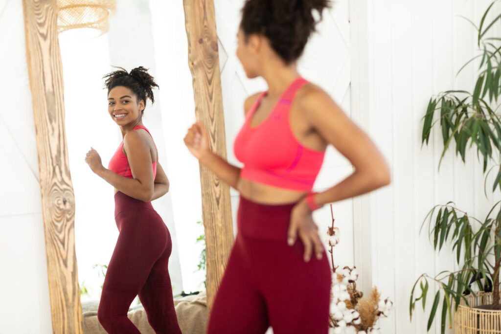 Joyful African woman after weight-loss looking in mirror standing indoors